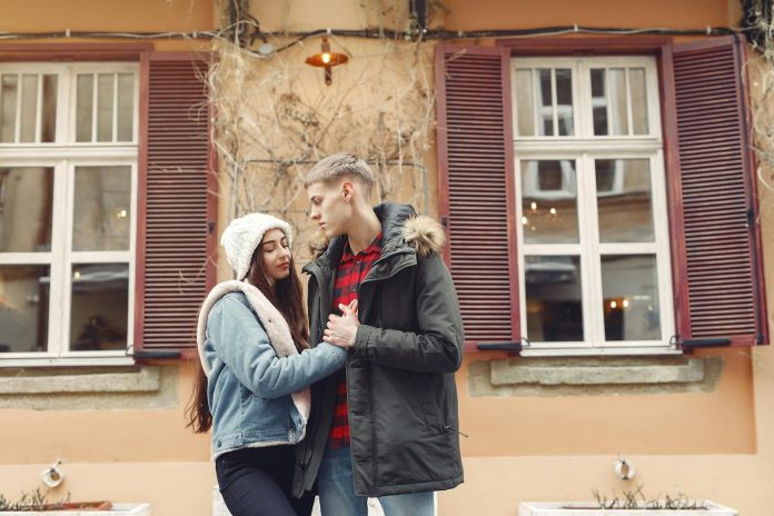 Young man and woman in warm clothes standing together on city street against old building and holding hands together during romantic date