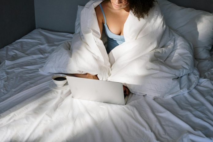 Woman cozily working from home, wrapped in a blanket with a laptop and coffee in bed.