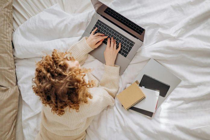 Woman with curly hair typing on a laptop in a cozy bedroom, surrounded by notebooks.